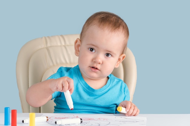 A baby of 12-17 months draws with felt-tip pens sitting on a child's chair