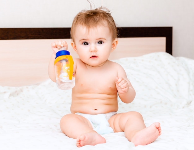 Il bambino di 6 mesi beve l'acqua da una bottiglia, sdraiato sul letto. bambino felice e gioioso