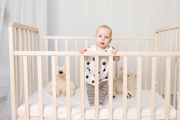 8 개월 된 아기가 밝은 어린이 방에서 잠옷을 입은 장난감을 가지고 침대에 서서 카메라를 바라본다