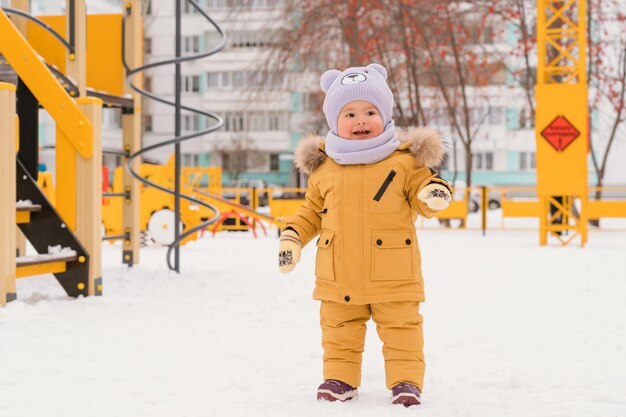 冬の遊び場で12〜17か月の赤ちゃん。黄色いジャケットを着た子供が、ロシアのアパートの中庭の雪の中で優しく立って微笑んでいます。