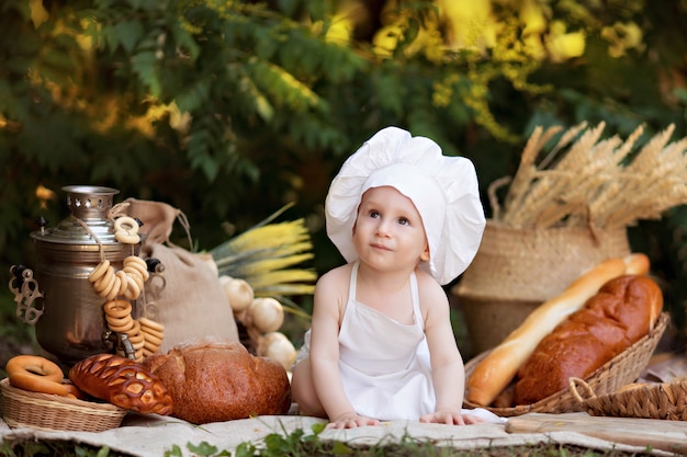 Il panettiere del bambino cucina in natura. il ragazzo su un picnic mangia pane e bagel in un grembiule bianco e cappello