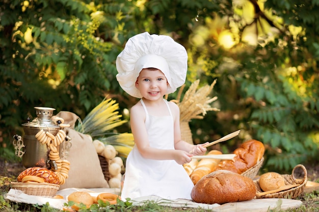 日当たりの良い夏の日にピクニックに赤ちゃんパン屋が白いエプロンでパンとベーグルを食べ、自然に帽子をかぶる