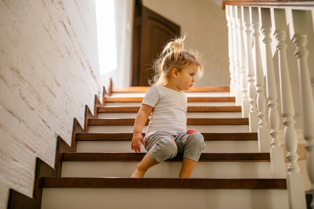 Baby blonde girl in white t-shirt at bottom of stairs indoors,looking at camera and smiling