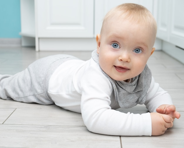 Baby blonde jongen met blauwe ogen in kleding van milieuvriendelijke materialen ligt op de vloer van het huis.