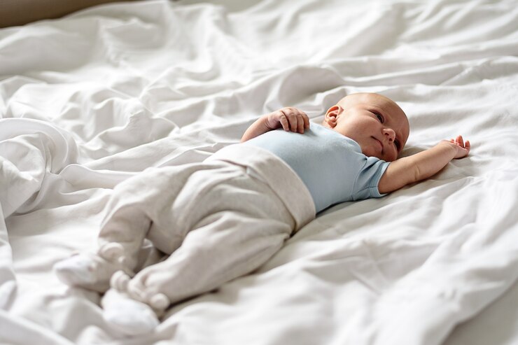 Baby in blue bodysuit and white pants lies with his open eyes on white bed