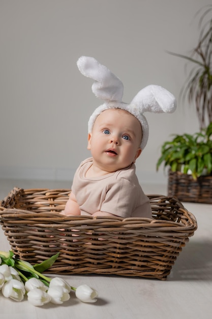 baby in bodysuit with rabbit ears is sitting in wicker basket on the floor with a bouquet of tulips