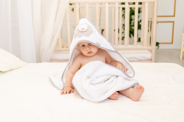 Photo baby boy 8 months old sitting on a white bed in a white towel after bathing in the bathroom at home