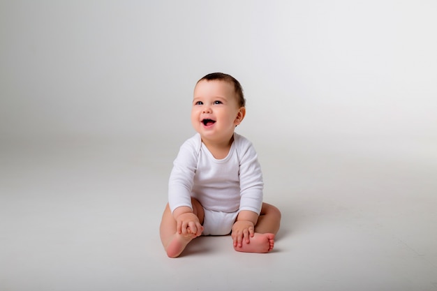 Bambino di 9 mesi in un body bianco seduto su un muro bianco