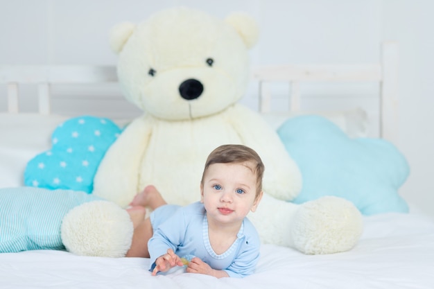 青いボディスーツの大きなテディベアとベッドの上の男の子、寝室で幸せな健康な小さな赤ちゃん