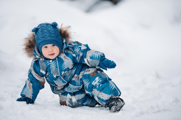파란 작업복을 입은 아기 소년이 깊은 눈 위에 앉아 아름다운 겨울날을 즐기고 있다
