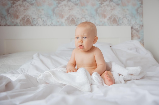 A baby boy in a diaper is sitting on the bed covered with a towel after bathing in the bedroom