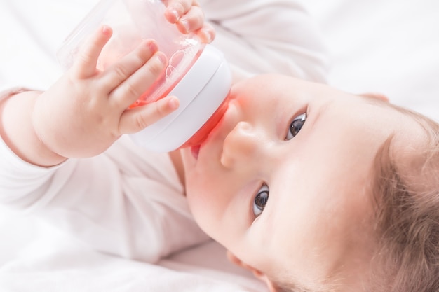 Baby boy lying on bed and drinking tea from her plastic bottle.