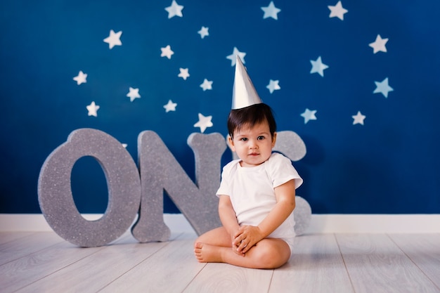 Baby boy of one year old wearing a white body and a festive paper hat sits on the floor on a blue background with stars and big silver letters one.