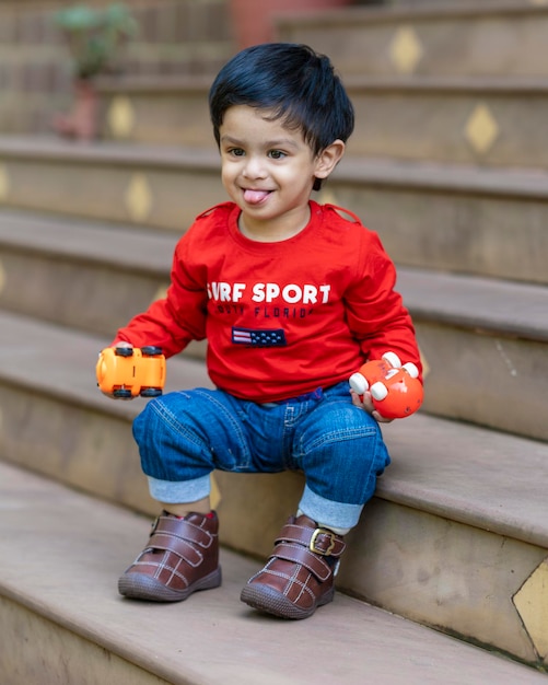 赤いTシャツとブルージーンズを着て階段に座っているおもちゃで遊ぶ男の子