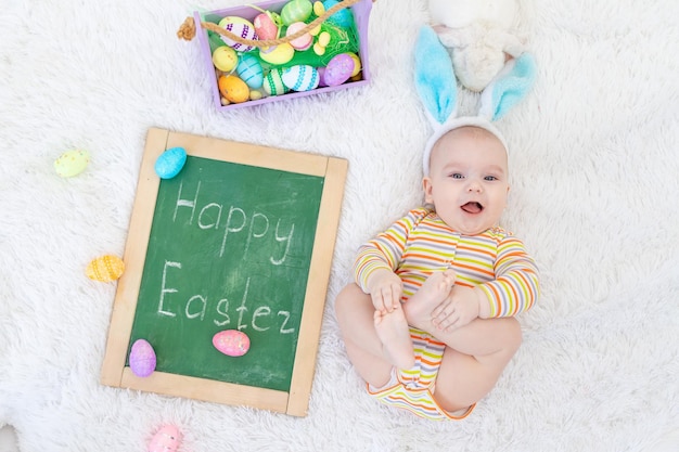 A baby boy in rabbit ears is lying on his head with Easter eggs and the inscription Happy Easter a cute funny smiling little baby The concept of Easter