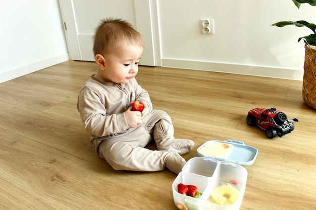 Photo a baby boy sits on the floor and eats food from a plastic tray