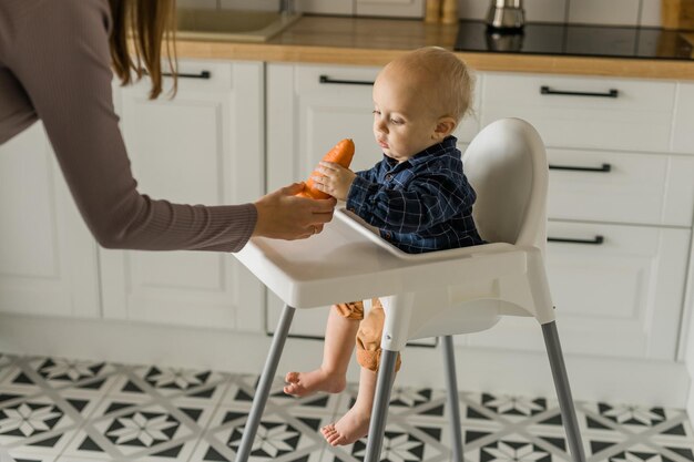 Baby boy sitting in baby chair eating carrot on kitchen background copy space baby feeding concept