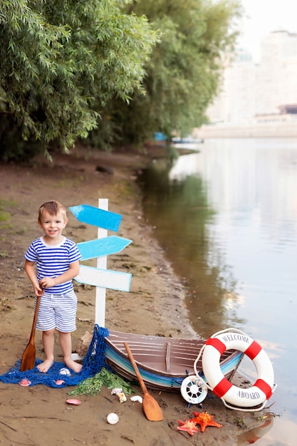 Baby boy sitting in a boat, dressed as a sailor, on a sandy beach with seashells by the sea