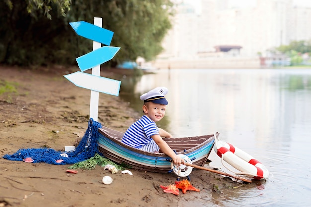 Photo baby boy sitting in a boat, dressed as a sailor on a sandy beach with seashells by the sea