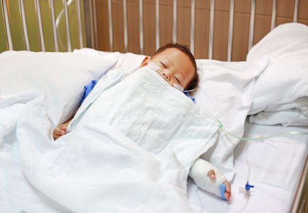 Baby boy sleeping with attaching intravenous tube to hand on bed at hospital.