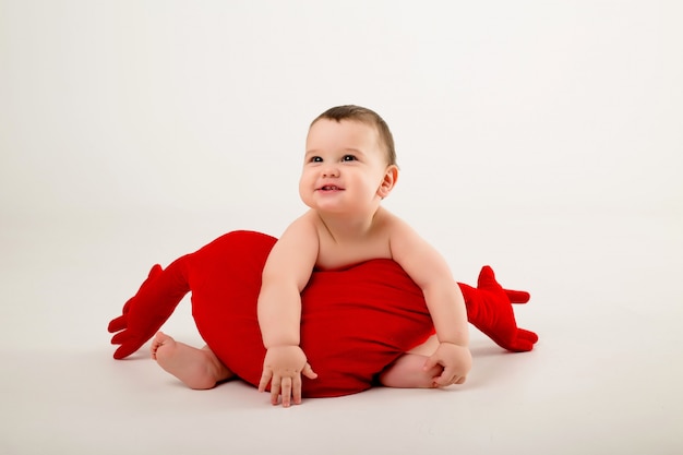 baby boy smiling and holding a red pillow in the shape of a heart, sitting on a white wall