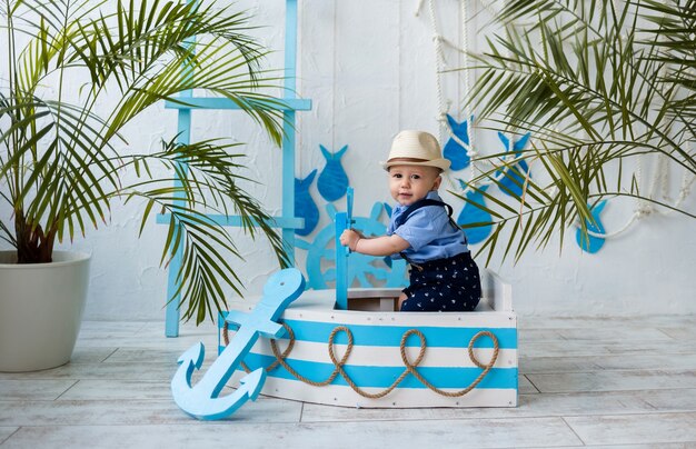 Baby boy in a straw hat sits in a white and blue wooden boat on a white surface with space for text