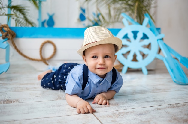 Baby boy in a suit and a straw hat lies on the wooden floor against a surface with a marine decor