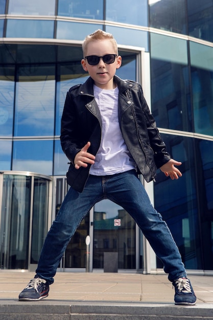 Photo baby boy in sunglasses and black leather jacket smiling on the background of the glass building