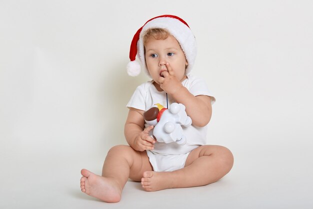 Baby boy wearing Santa Claus hat and white wall, playing with plastic dog toy, looking away, biting his fingers, sitting barefoot on floor, looking away.