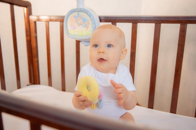 A baby boy in a white bodysuit is sitting in a crib drinking\
smile