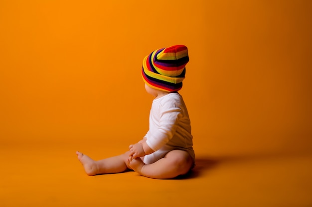 Photo baby boy in a white bodysuit and multicolored hat holding a toy sitting on a yellow wall