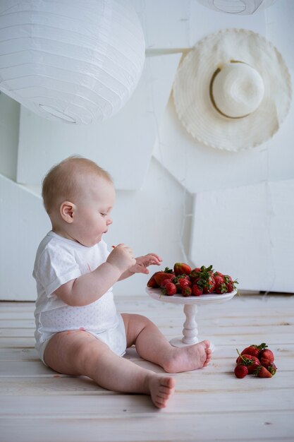 Baby boy in a white bodysuit sits with a plate of strawberries on a white wall with space for text