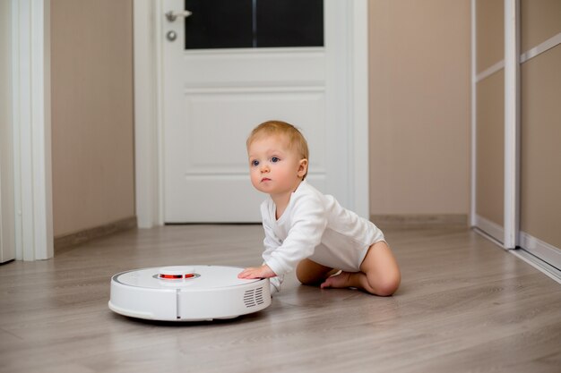 baby boy in white clothes with a robotic vacuum cleaner on the floor of the house