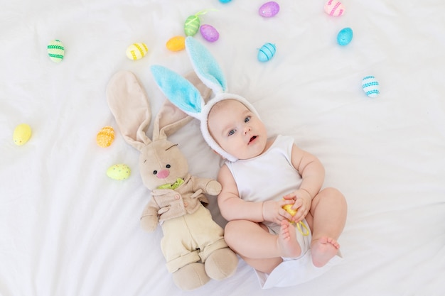 A baby boy with rabbit ears on his head is lying in a crib with a bunny toy and Easter eggs, a cute funny smiling little baby. Easter Concept