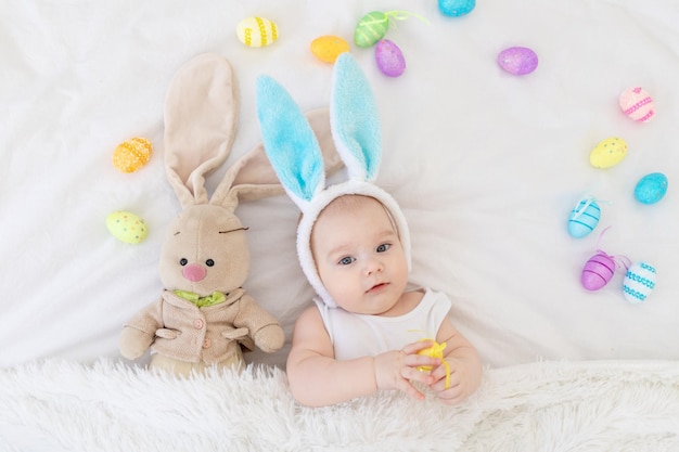 A baby boy with rabbit ears on his head is lying in a crib with a bunny toy and Easter eggs a cute funny smiling little baby Easter Concept