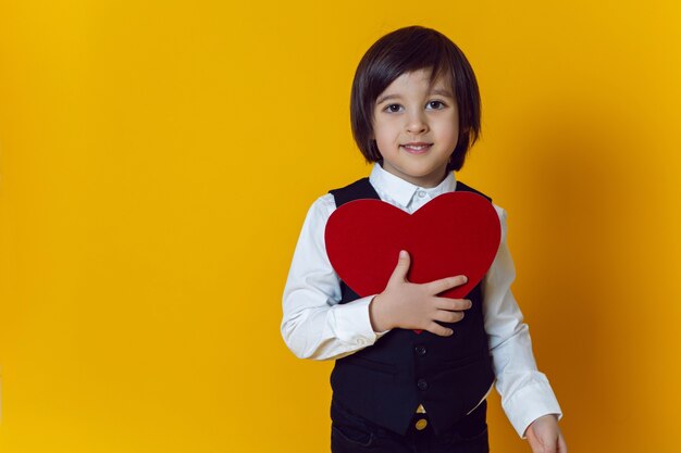 Baby boy with red paper heart in costume stands in yellow wall on valentine's day