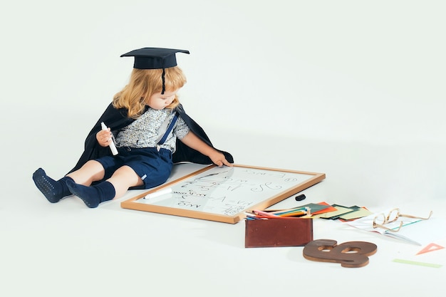 Baby child in academic gown and hat drawing by marker on blackboard near school supplies on white  isolated