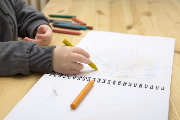 A baby child draws with colorful crayons in an album on a wooden table