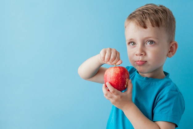 Baby child holding and eating red apple on blue wall, food, diet and healthy eating concept.