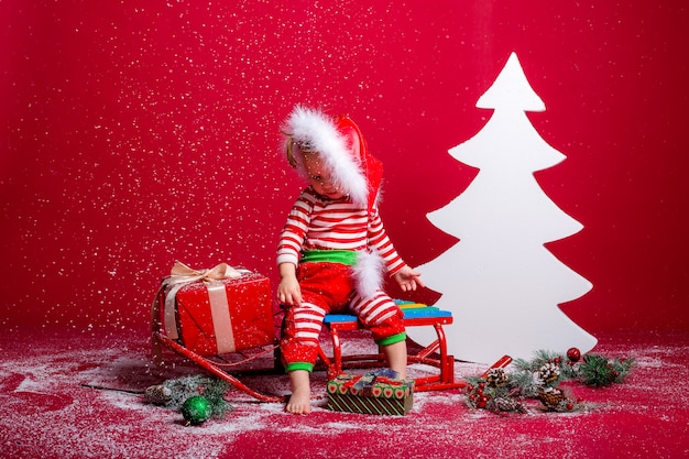 baby in Christmas pajamas and Santa hat catches snow sitting on a sled with gift box and big white Christmas tree on red background