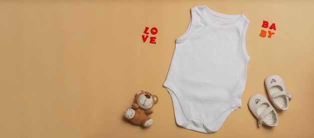 Фото Белый комбинезон для новорожденных обуви и плюшевый медведь на бежевом фоне пространство для текста верхний вид баннер