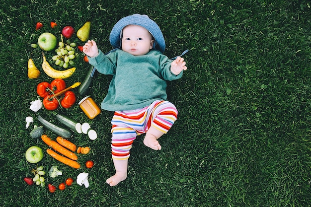 다채로운 옷을 입은 아기가 음식을 시도하고 푸른 잔디에 다양한 신선한 과일 채소를 틀고 있습니다