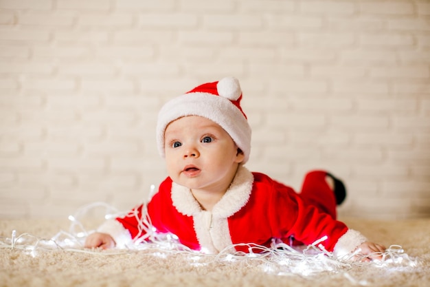 The baby in costume Santa Claus lies on the floor with garlands