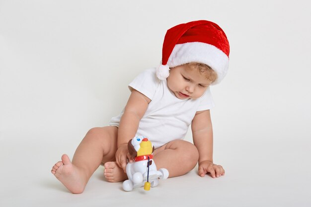 Baby draagt bodysuit en kerstmuts spelen met nieuwe speelgoed witte blootsvoets zitten geïsoleerd over witte ruimte