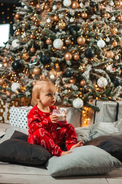 A baby drinks milk in a glass near the tree at Christmas