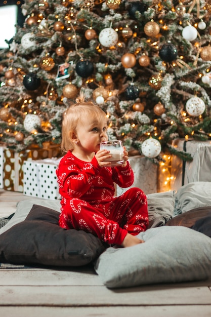 A baby drinks milk in a glass of Santa Claus at Christmas near the tree
