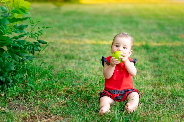 A baby eats a green apple in a red bodysuit on the green grass in summer, space for text
