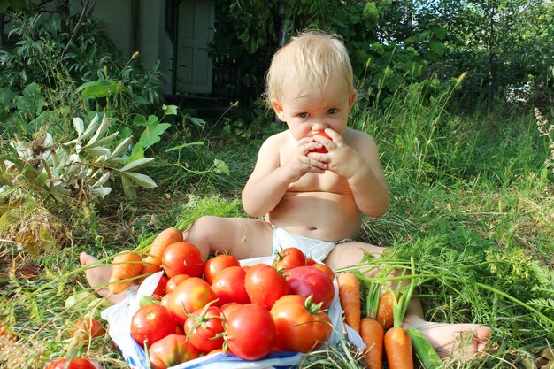 baby eet rijpe tomaat naast een hoop tomaten en wortelen