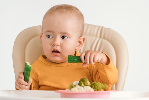 Ребенок на стульчике для кормления двумя руками ест свежий зеленый огурец. На белом фоне.