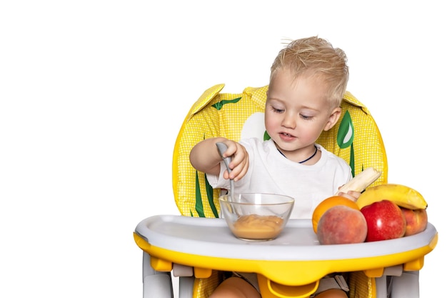 Baby feeding A little boy eats fruit puree with a spoon by himself sitting in a child seat in the kitchen Closeup portrait